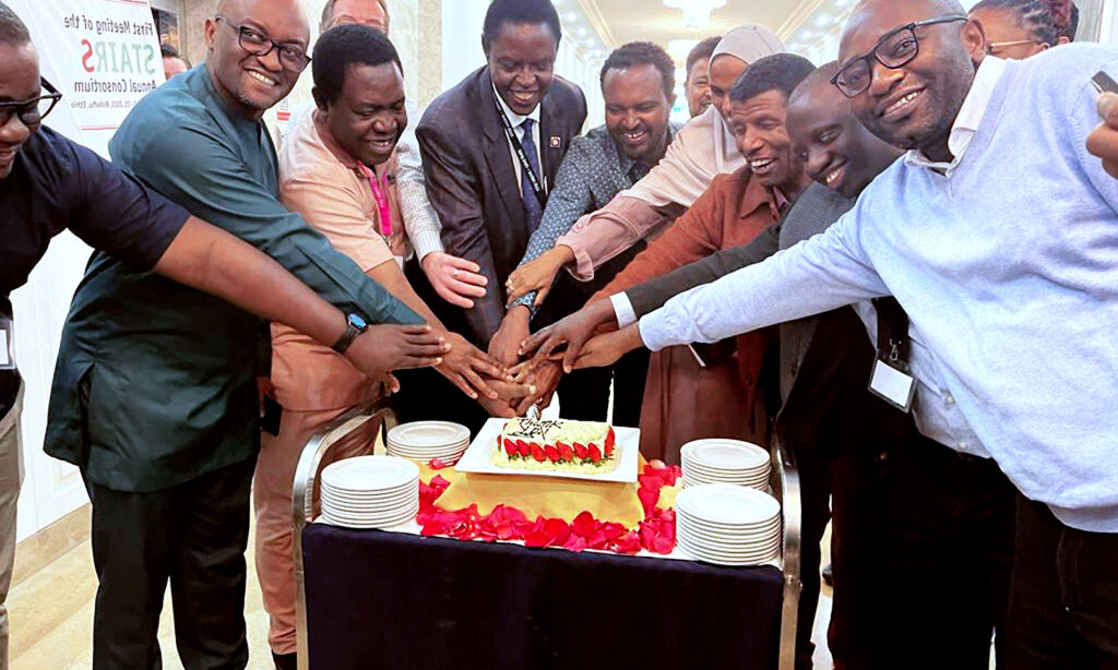 Group of people cheering and making a collaborative team gesture with their hands over a symbolic cake standing in a foyer.